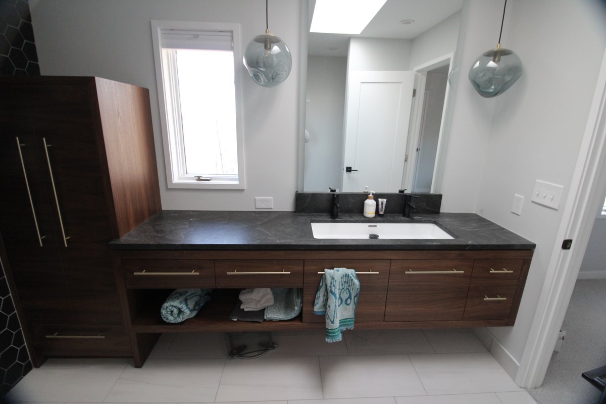 Walnut bathroom vanity with a dark counter, glass pendant lights and a tall storage cabinet.