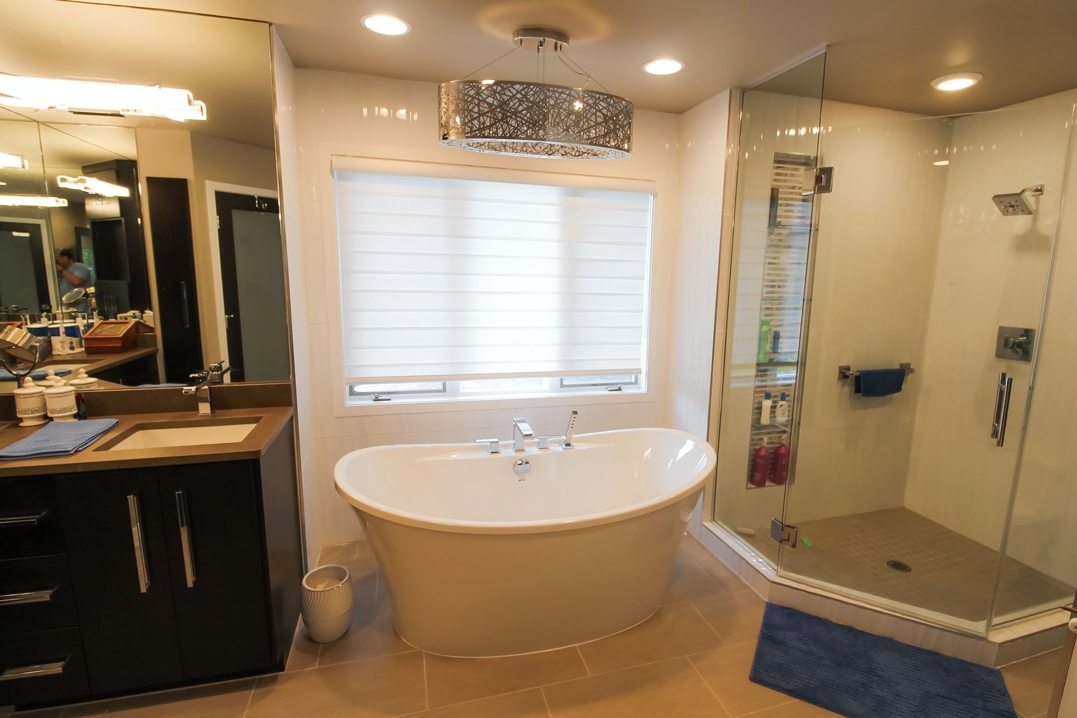 Freestanding tub under a drum chandelier with glass shower and dark vanity